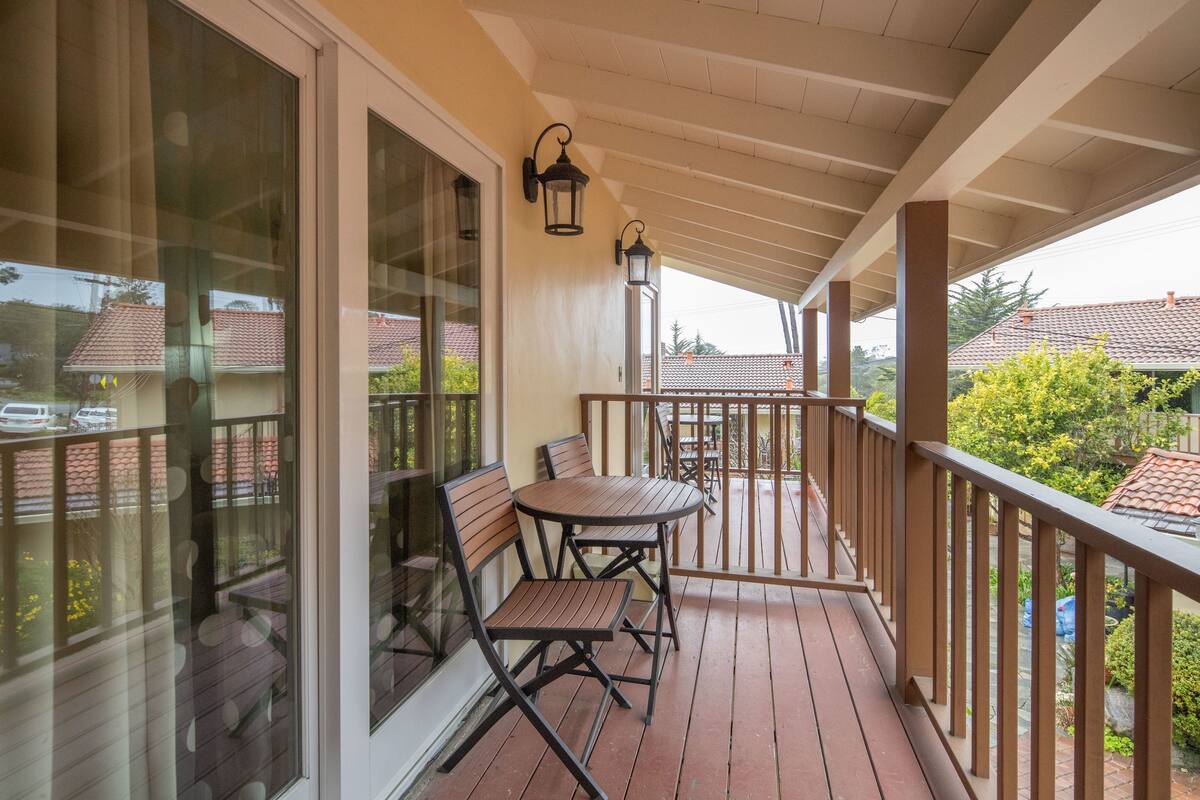 A covered balcony with a wooden table and chairs, large windows, and a view of neighboring houses.