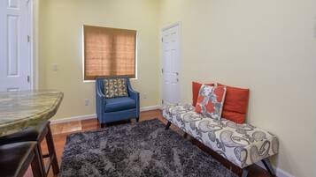 Um quarto com uma mesa de jantar, uma poltrona azul, um sofá com almofadas estampadas e uma grande janela com persianas.