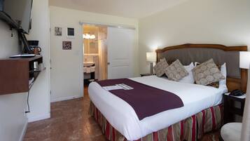 A hotel room with a large bed, a desk with a computer, a chair, a bedside table with a lamp, and a bathroom visible through an open door.