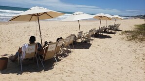 Ein Strand mit Reihen von Liegestühlen und Sonnenschirmen, eine Person sitzt auf einem Liegestuhl, und im Hintergrund das Meer.