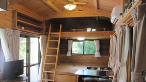 A wooden cabin interior with a dining table, benches, a ladder, a television on a stand, and a window with curtains.