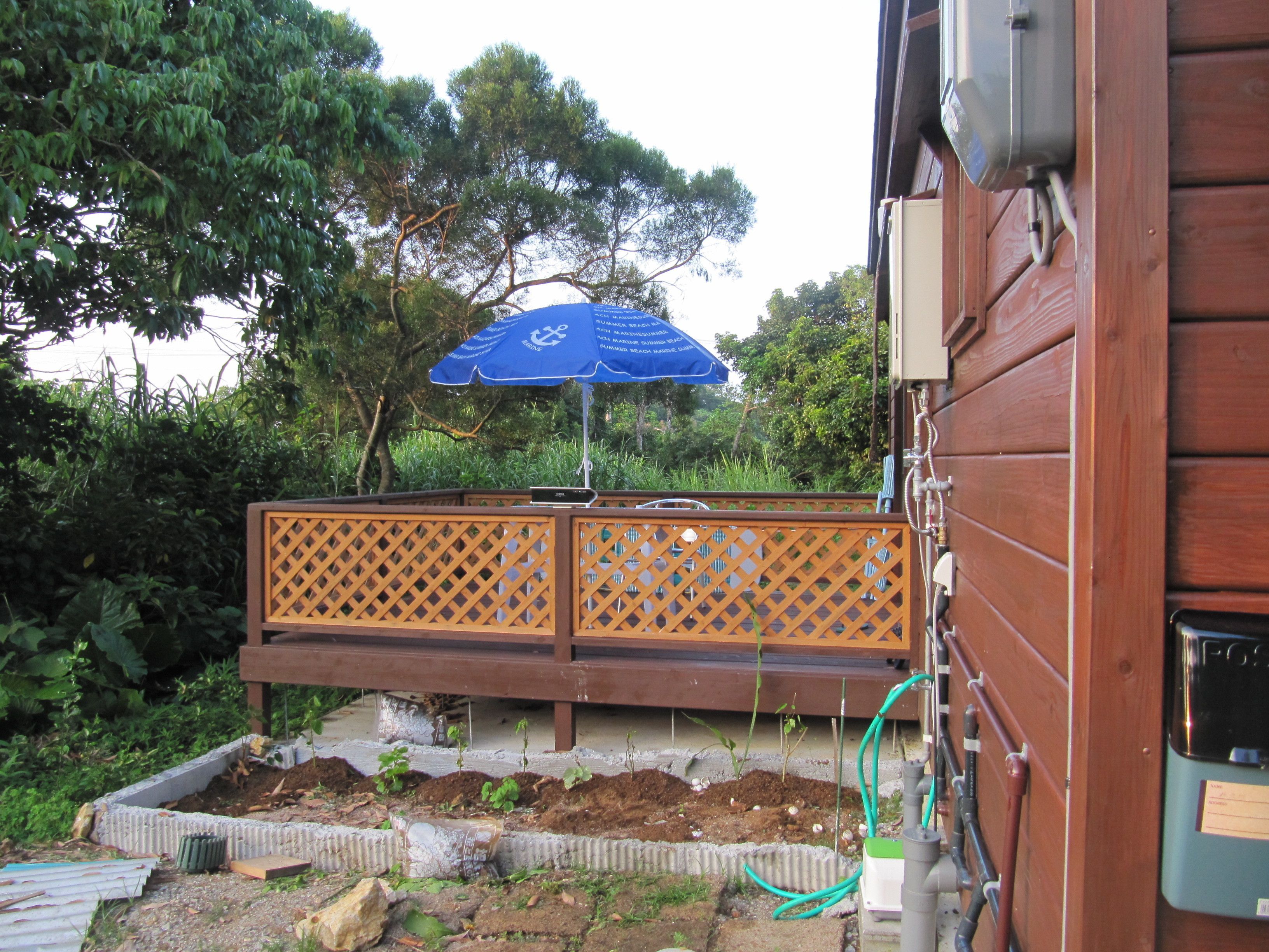 Una terraza de madera con una sombrilla azul, rodeada de abundante vegetación y una cerca de madera.