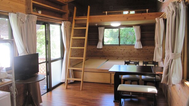A wooden cabin interior with a wooden table, chairs, a bench, a TV, a ladder, and a window with curtains.