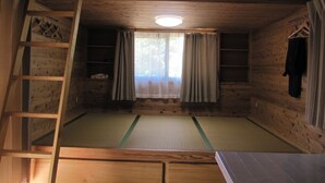 A wooden room with a tatami mat floor, a ladder, a window with curtains, and a shelf with items.