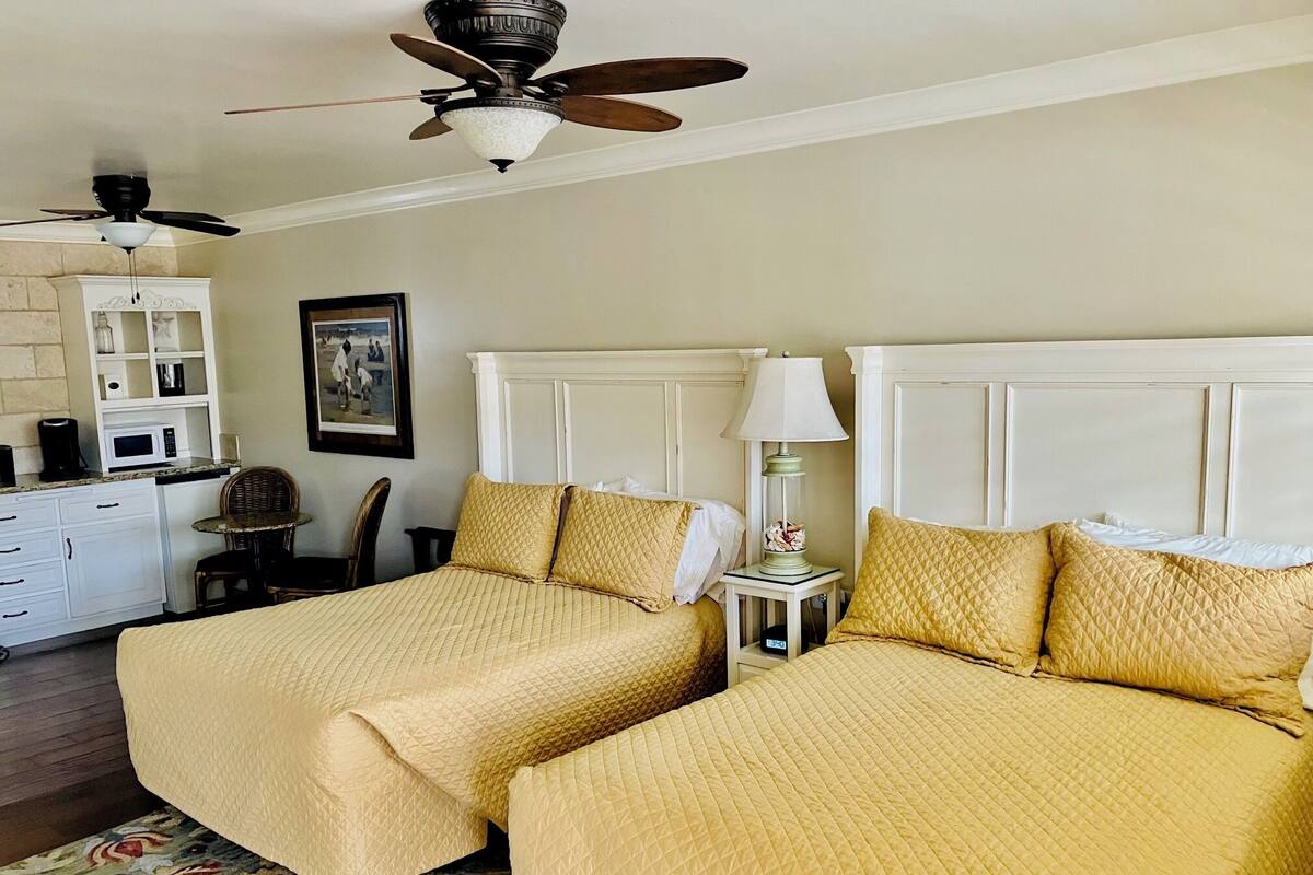 A hotel room with two beds, a ceiling fan, a microwave, a small table with a lamp, and a chair.