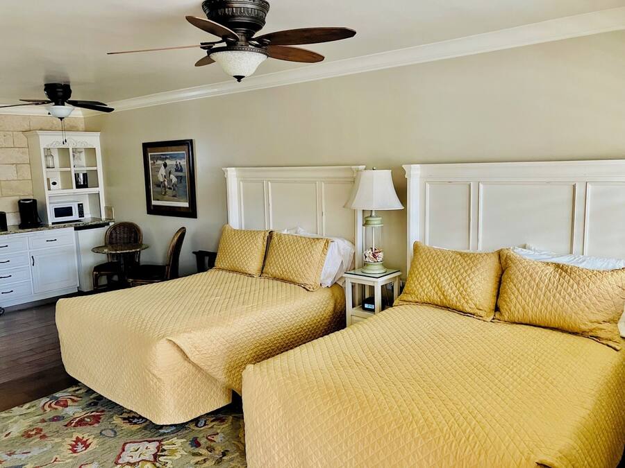 A hotel room with two beds, a ceiling fan, a microwave, a small table with a lamp, and a chair.