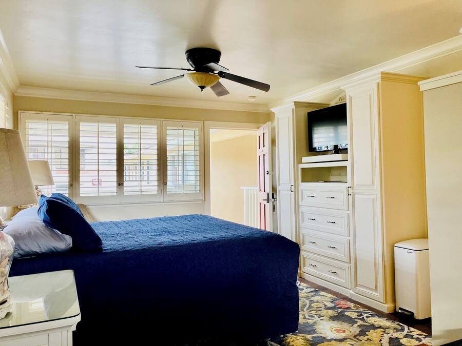 Un dormitorio con una cama, ventilador de techo, televisor sobre un mueble y una ventana con persianas.