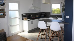 A modern kitchen with white cabinets, a central island with bar stools, a window with a view, and a hanging light fixture. - A haven of peace with Spa at 2 steps from the beach (Cléder)