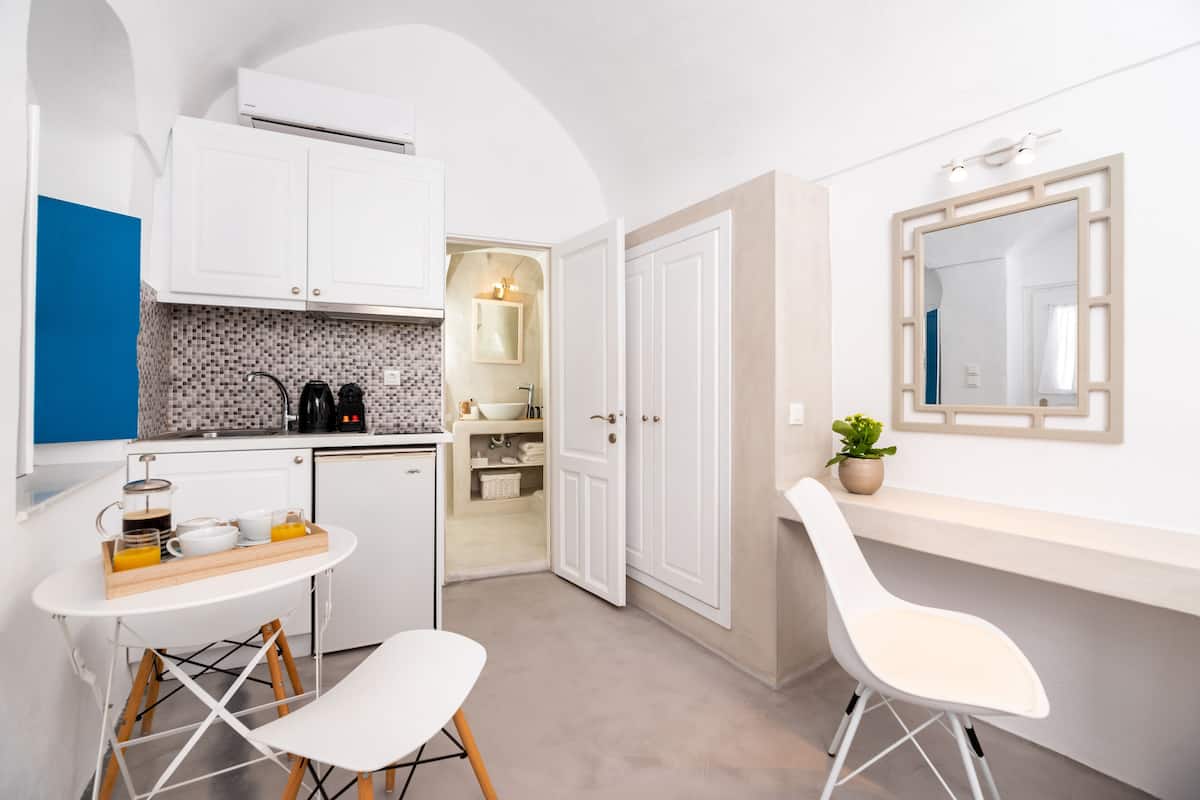 A compact kitchen with white cabinets, a coffee maker, and a breakfast setup. There is a white chair and a small table with a tray of orange juice.