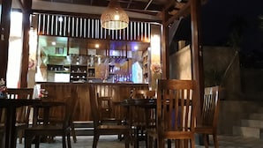 An outdoor dining area with wooden tables and chairs, a bar with shelves of bottles, and a hanging light fixture. - Grand Raka Homestay (Buleleng)