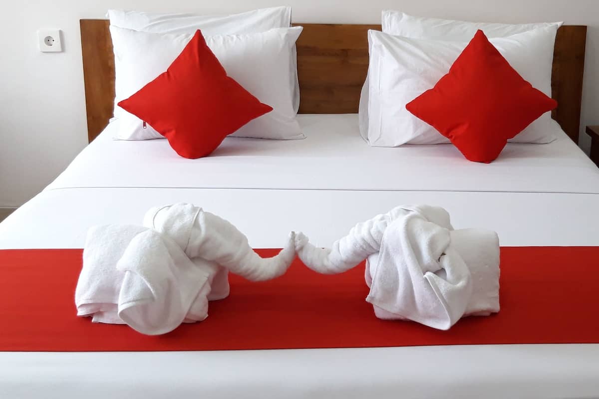 A hotel room with a bed featuring white bedding and two red pillows, a wooden headboard, a bedside table with a lamp, and two towels arranged on the bed.