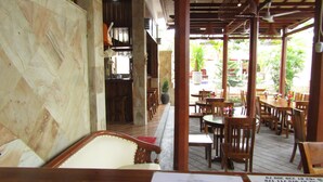A restaurant with wooden tables and chairs, a stone wall, and a view of greenery outside. - Grand Raka Homestay (Buleleng)
