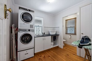 A laundry room with stacked washers and dryers, a sink, a window with blinds, and an ironing board with an iron. - Frangipani House (East Toowoomba)