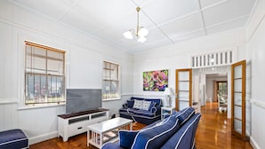 A spacious living room with a large TV, blue sofas, wooden flooring, and white paneled walls. - Frangipani House (East Toowoomba)