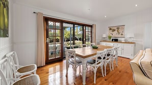 A dining area with a wooden table and white chairs, a kitchen with white cabinets, and a view through large glass doors. - Frangipani House (East Toowoomba)