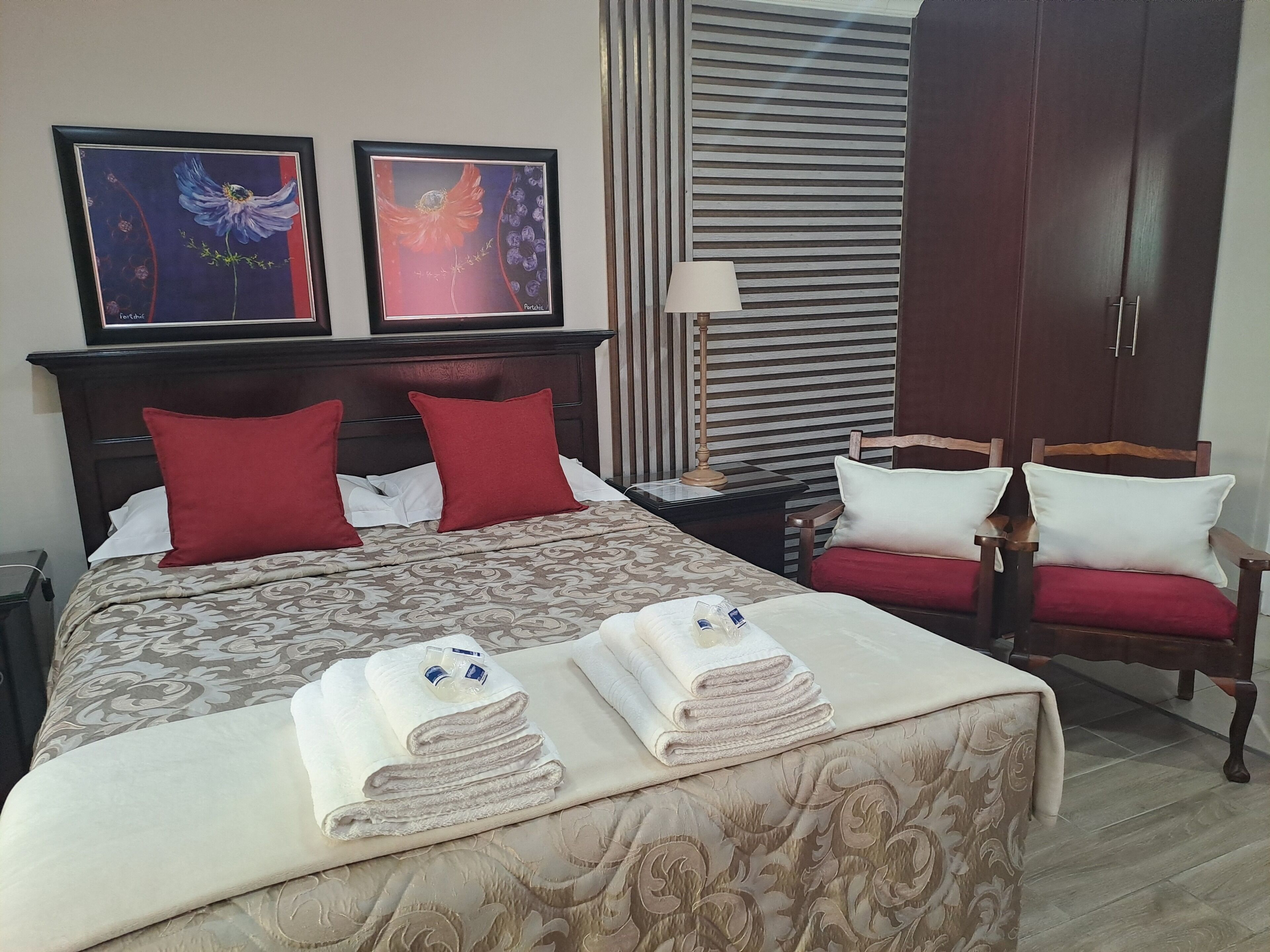 A hotel room with a large bed, two red pillows, a wooden headboard, a nightstand with a lamp, a wardrobe, and two framed artworks on the wall.