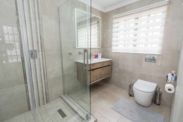 A modern bathroom with a glass shower enclosure, a wooden vanity, a toilet, and a window with blinds.