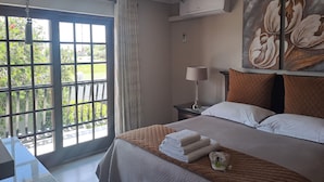 A hotel room with a large window, a bed with neatly folded towels, a nightstand with a lamp, and a wall-mounted air conditioner. - Kuiersaam Gastehuis (Secunda)