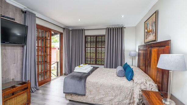 A hotel room with a bed, a TV, a wooden headboard, a lamp, and a window with curtains. - Kuiersaam Gastehuis (Secunda)