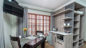 A dining area with a table set for two, a TV mounted above the table, and a kitchen area with wooden cabinets and shelves. - Kuiersaam Gastehuis (Secunda)