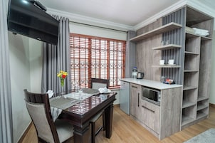 Un comedor con una mesa dispuesta para dos, un televisor montado sobre la mesa, y una zona de cocina con armarios y estantes de madera.