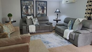 A living room with two grey sofas, a coffee table, and a floor lamp. There are two framed pictures on the wall, a potted plant, and a small table with decorative items. - Kuiersaam Gastehuis (Secunda)