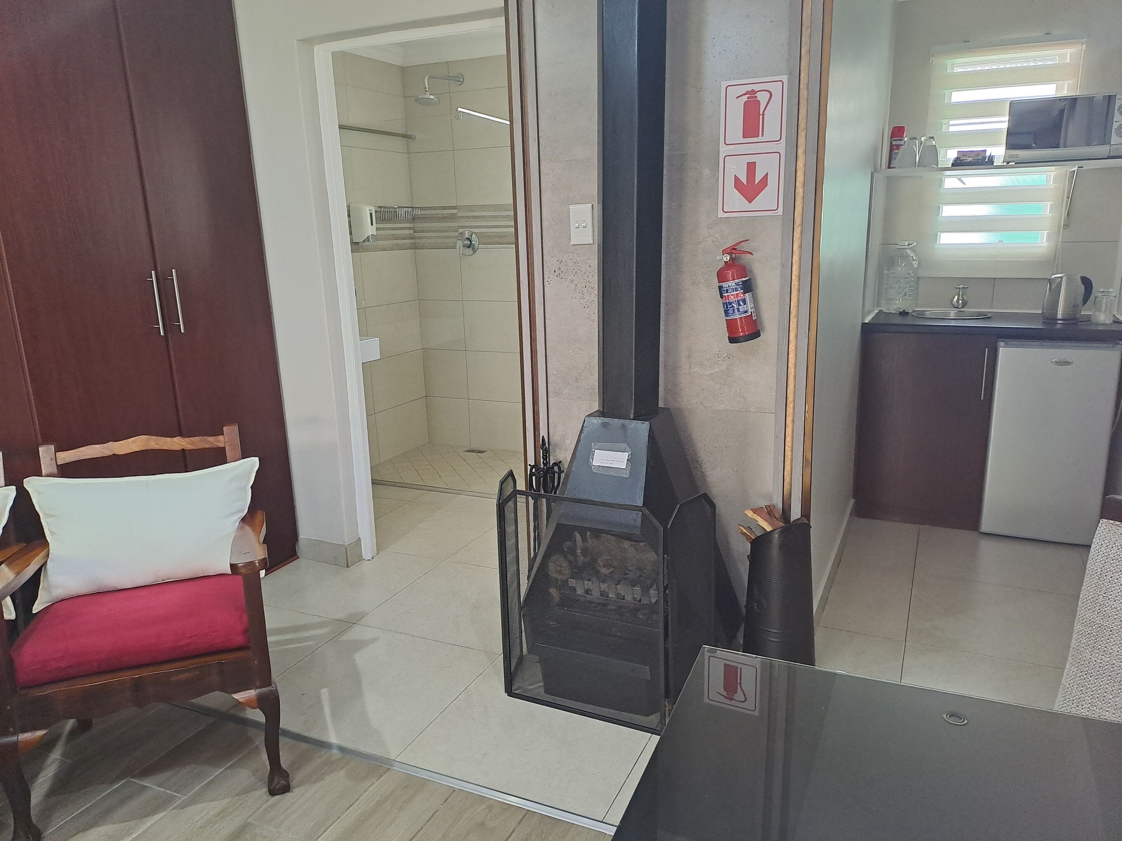 A hotel room with a wooden wardrobe, a chair with a red cushion, a glass partition, a kitchenette with a refrigerator, a microwave, and a fire extinguisher.