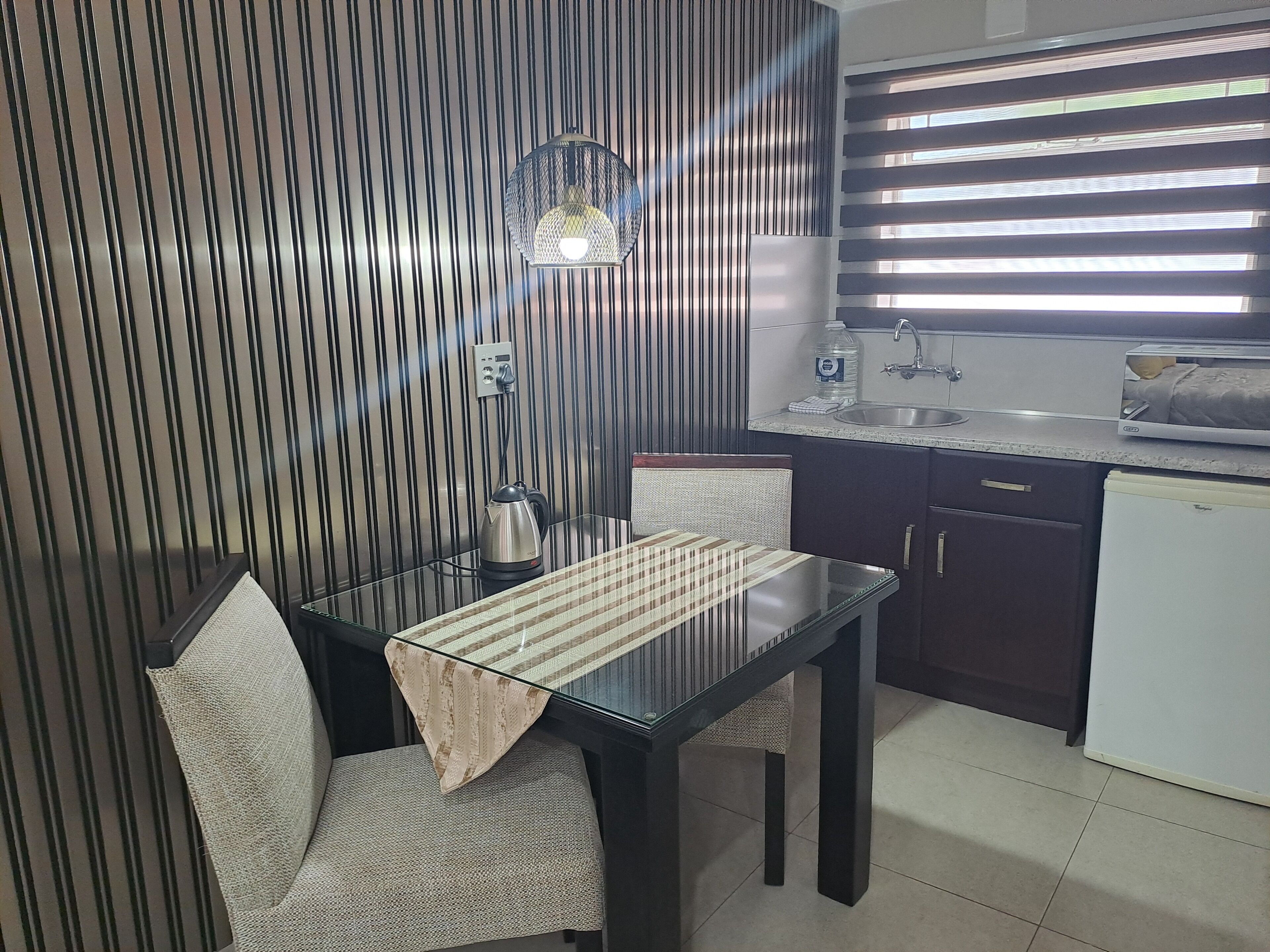 A compact kitchen with a dining table, chairs, a kettle, and a refrigerator.