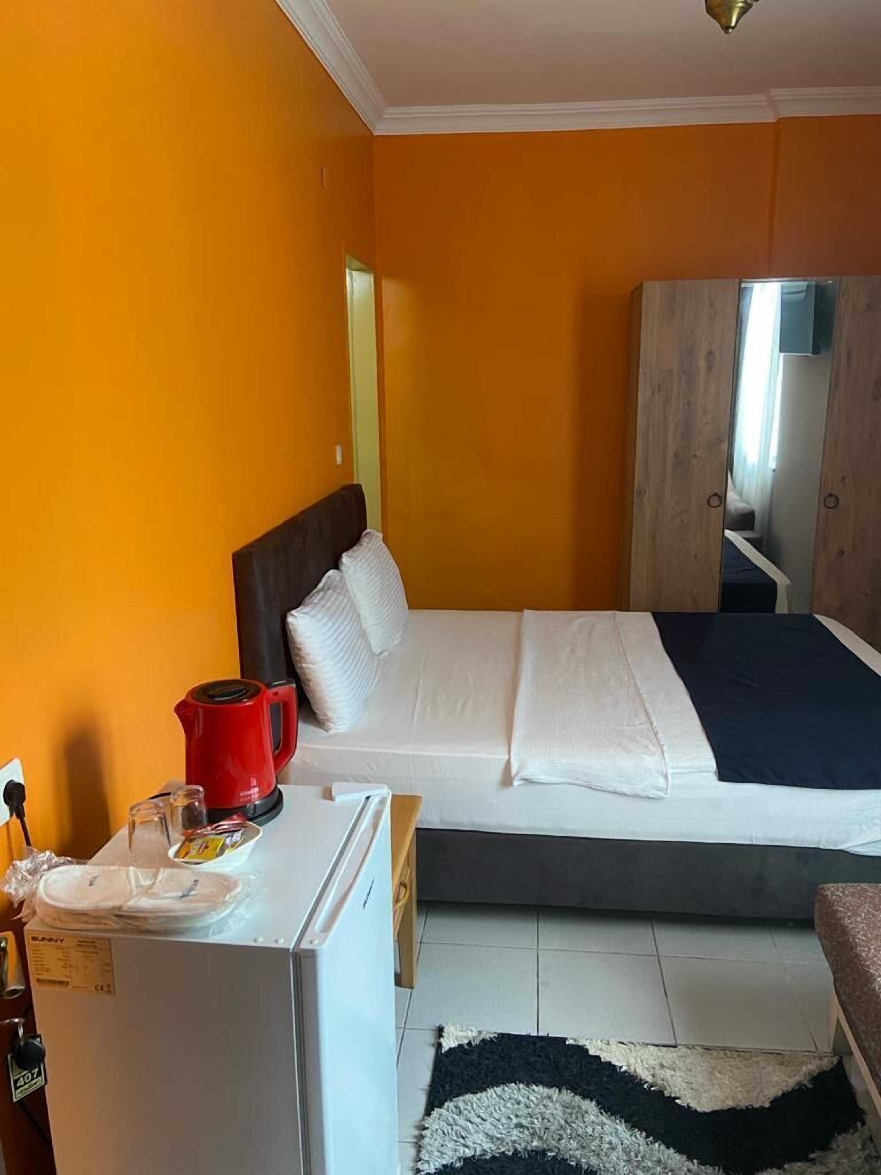 A bedroom with a bed, a small refrigerator, a red kettle, and a wooden wardrobe.