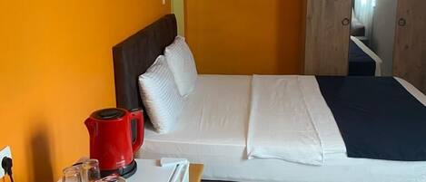 A bedroom with a bed, a small refrigerator, a red kettle, and a wooden wardrobe.