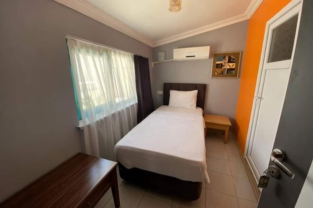 A bedroom with a bed, bedside table, window with sheer curtains, and a door.