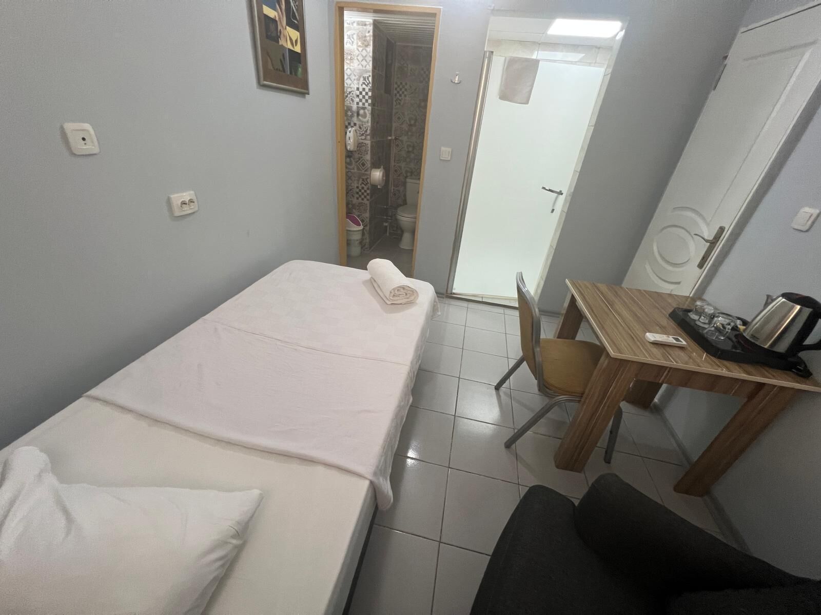 A small hotel room with a single bed, a wooden table with a kettle and glasses, a chair, and a door leading to a bathroom.