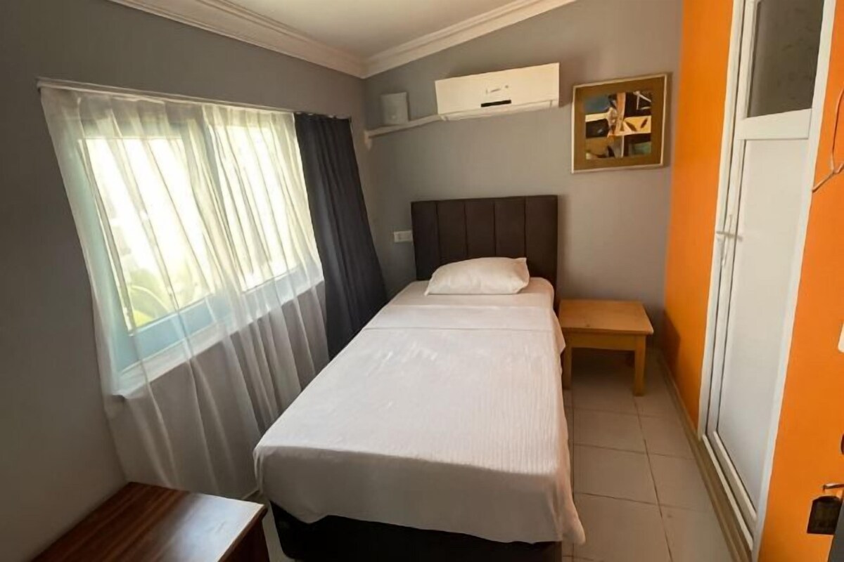 A room with a bed, a bedside table, a window with curtains, a wall-mounted air conditioner, and a small shelf with a picture.