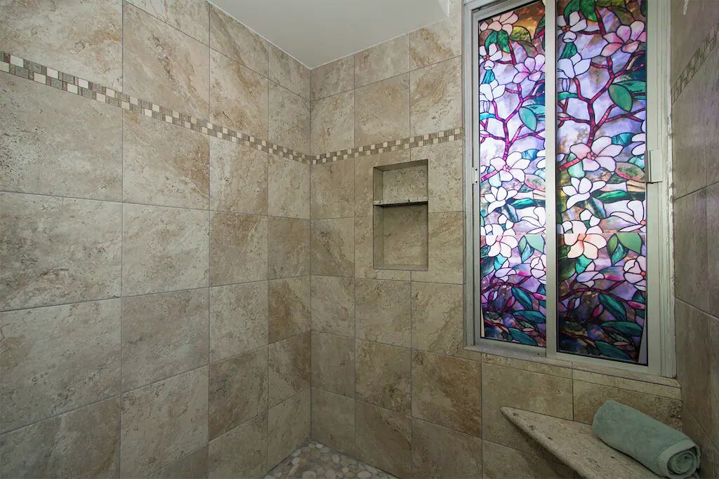 A shower with beige tiles, a small window with stained glass, and a towel on the floor.