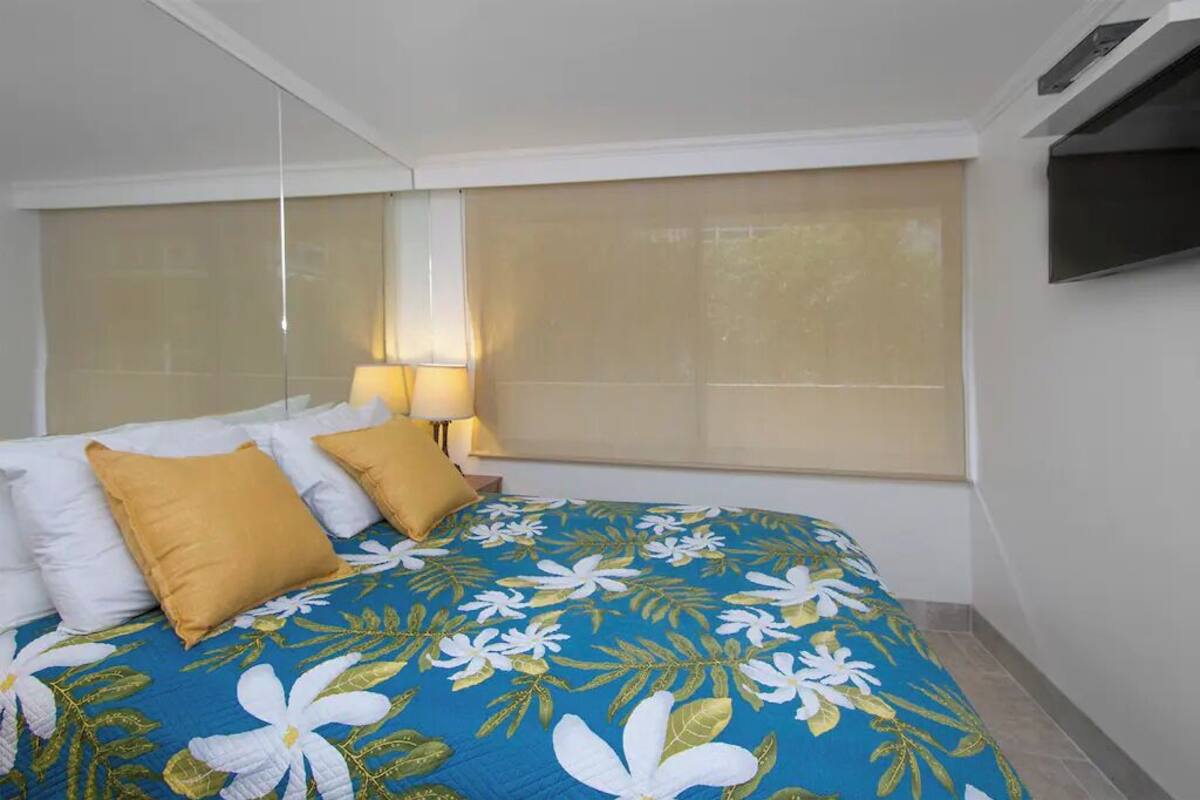 A hotel room with a large bed, a TV mounted on the wall, and a window with blinds.