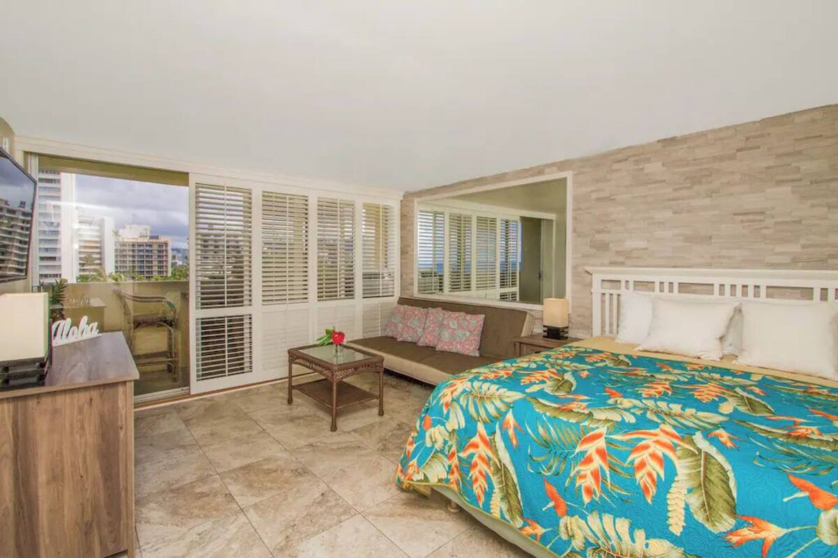 A hotel room with a bed, a sofa, a desk, and a large window with blinds.