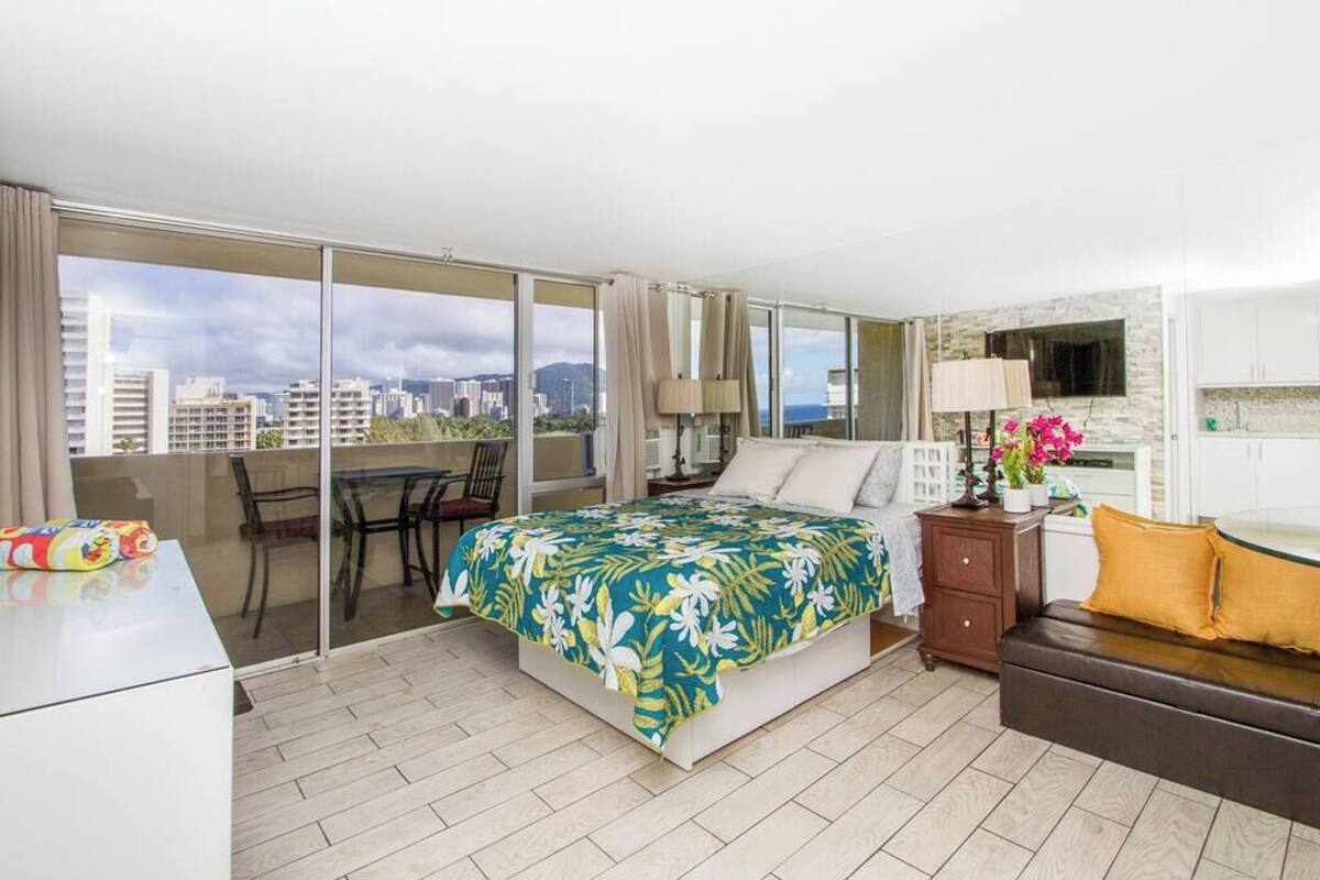 A hotel room with a bed, a desk, a chair, a sofa, a TV, and a balcony with a city view.