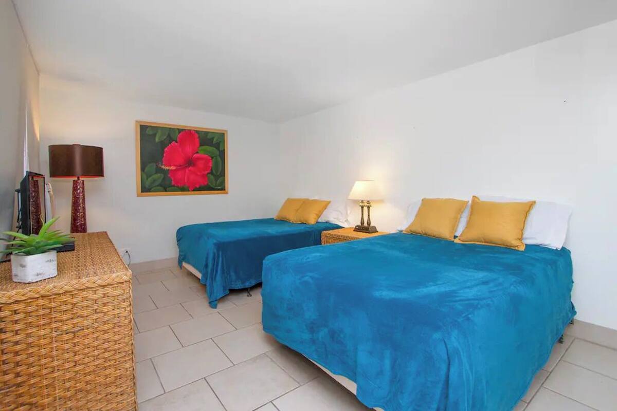 A room with two beds, each with blue bedding and yellow pillows. There is a painting of a red flower on the wall, a lamp on each side of the beds, and a wicker side table with a plant.