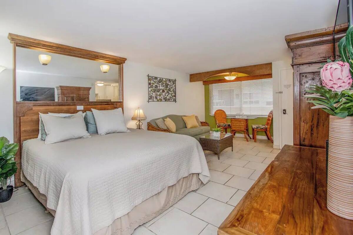 A bedroom with a large bed, a wooden headboard, a mirror, a green wall, a window with blinds, a small table with a lamp, and a wooden cabinet with a plant.
