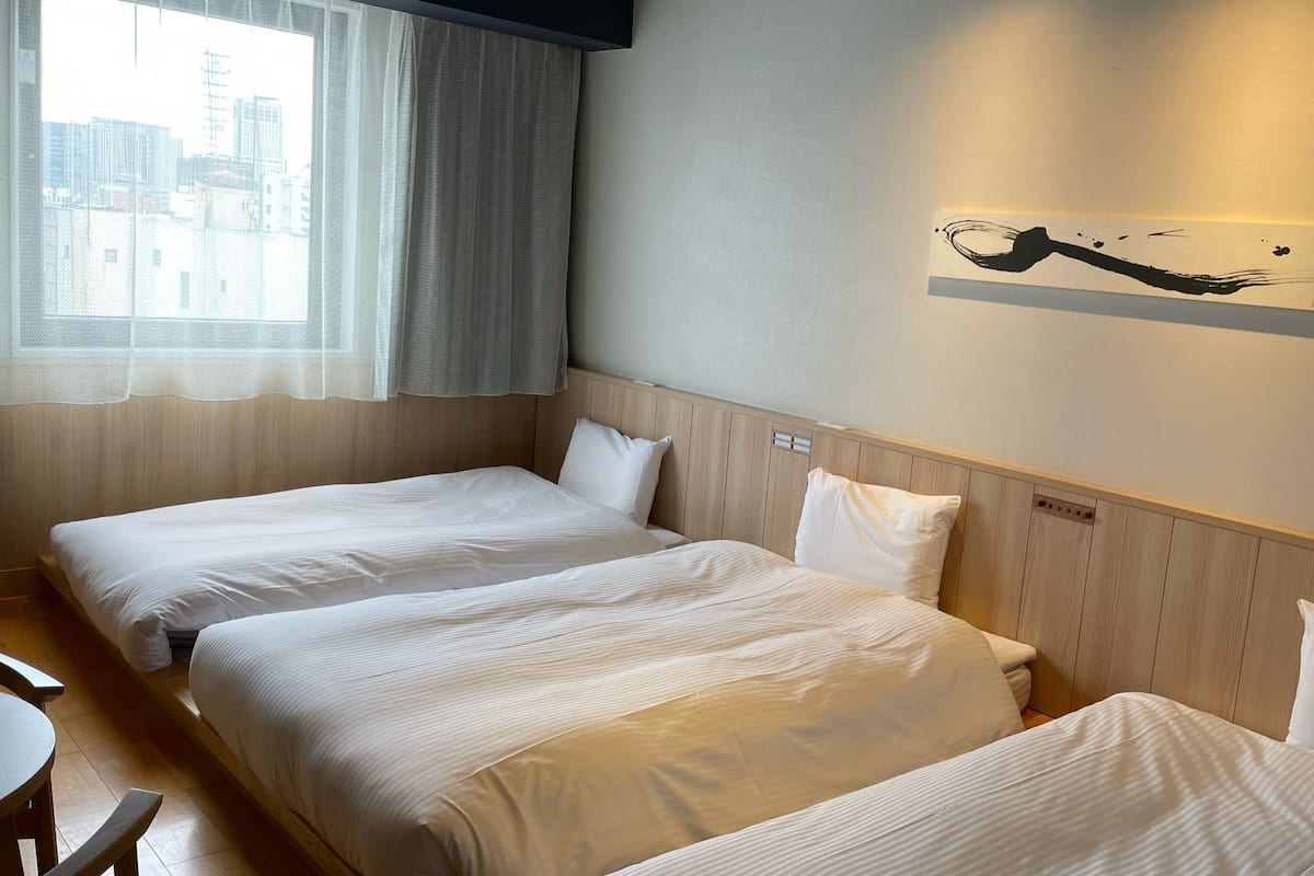 A hotel room with two single beds, a wooden headboard, a city view through a window with sheer curtains, and a wall-mounted artwork.
