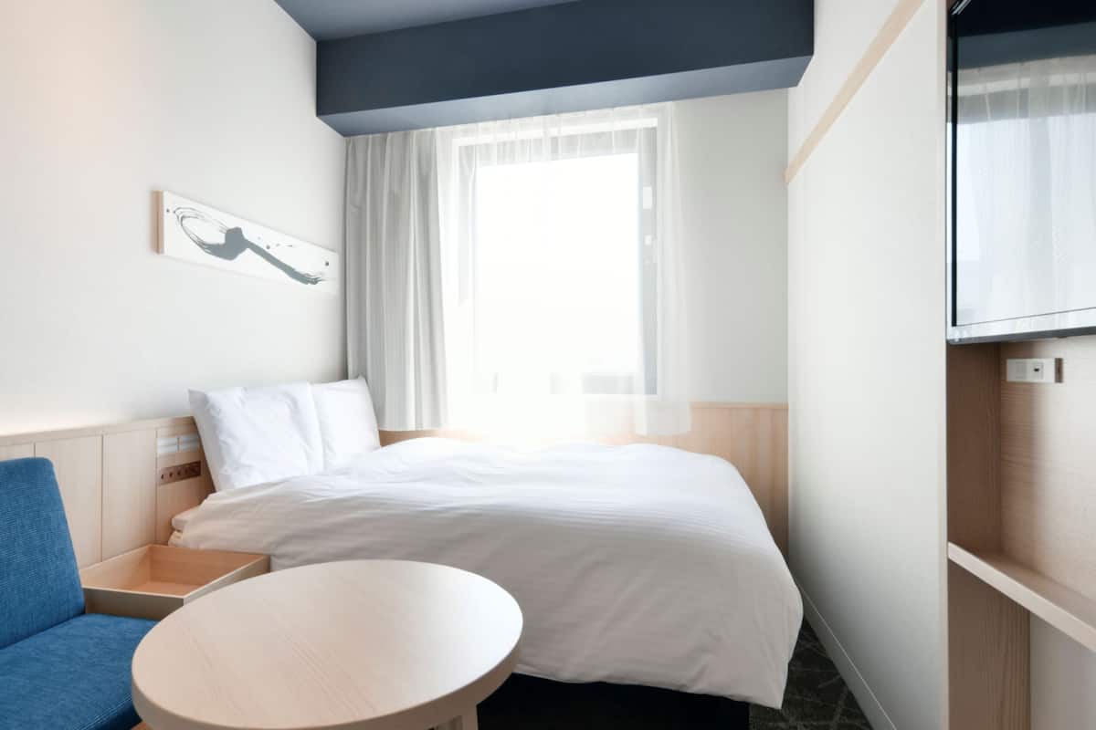 A hotel room with a bed, a blue sofa, a small round table, a painting on the wall, and a TV mounted on the wall.