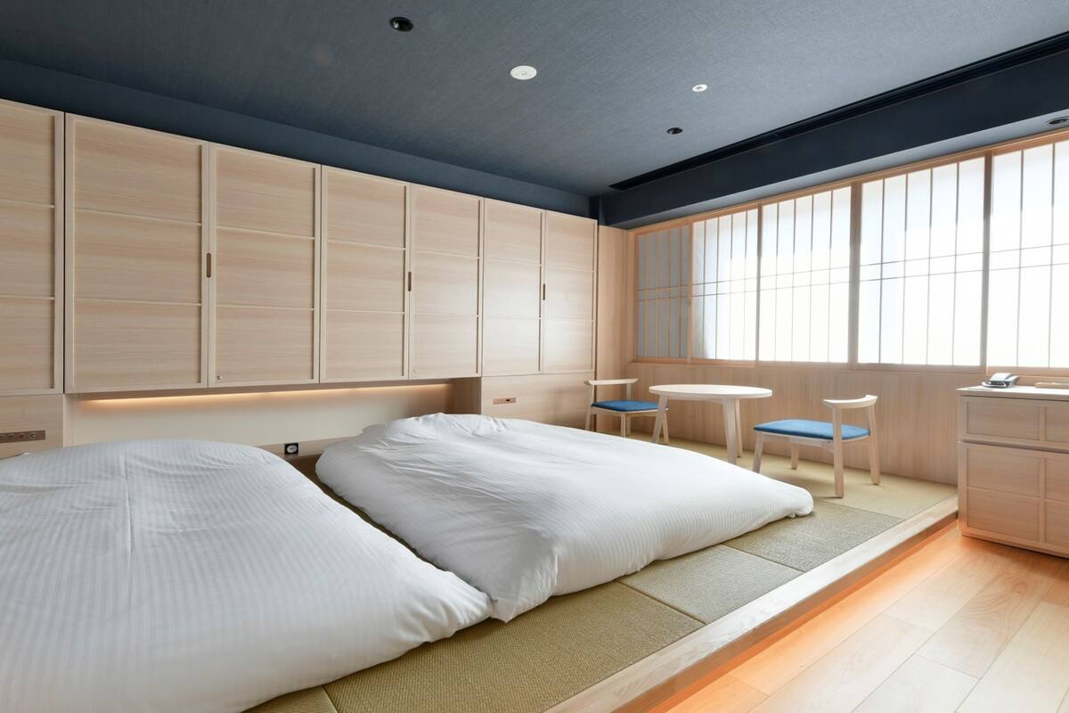 A modern bedroom with two beds, a small table, and chairs, and large windows providing natural light.