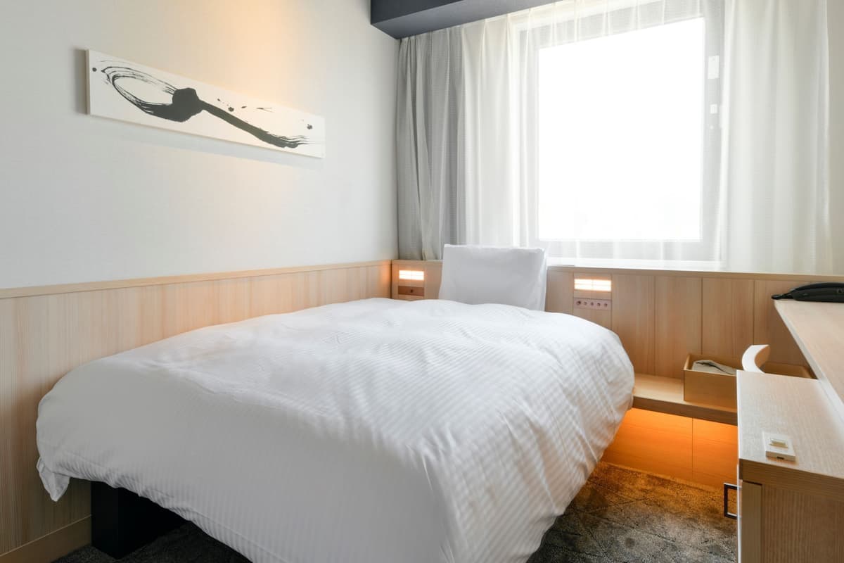 A hotel room with a bed, a desk, a chair, a nightstand, and a window with curtains.