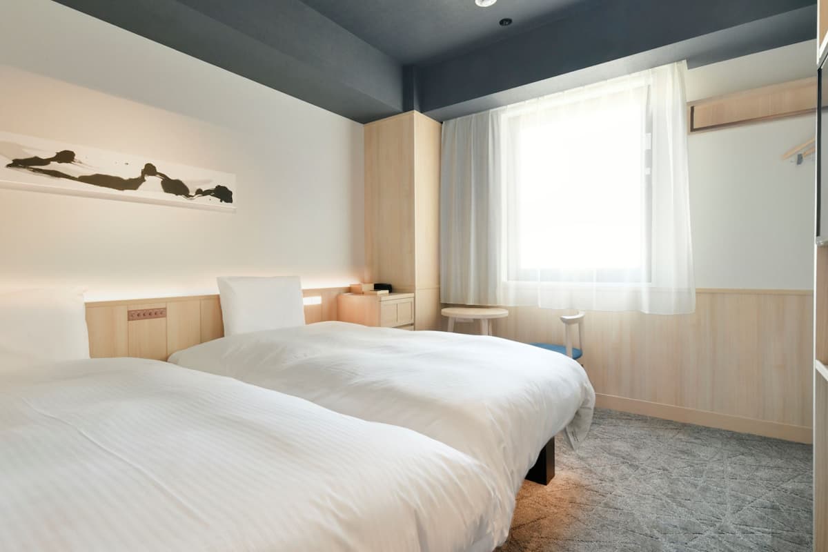 A hotel room with a large bed, a bedside table, a small stool, a window with curtains, and a wall-mounted artwork.