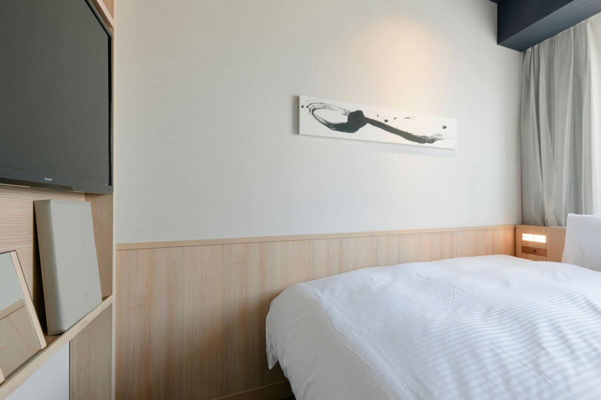 A hotel room with a bed, a mounted flat-screen TV, a wall-mounted artwork, and a window with curtains.