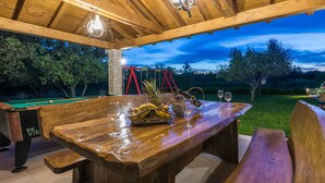 Een houten terras buiten met een pooltafel, een biljarttafel en een eettafel gedekt met fruit en glazen.