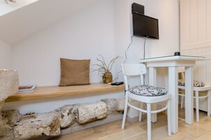 A cozy room with a stone wall, a wooden bench, a white table with chairs, a mounted TV, and a vase with branches.