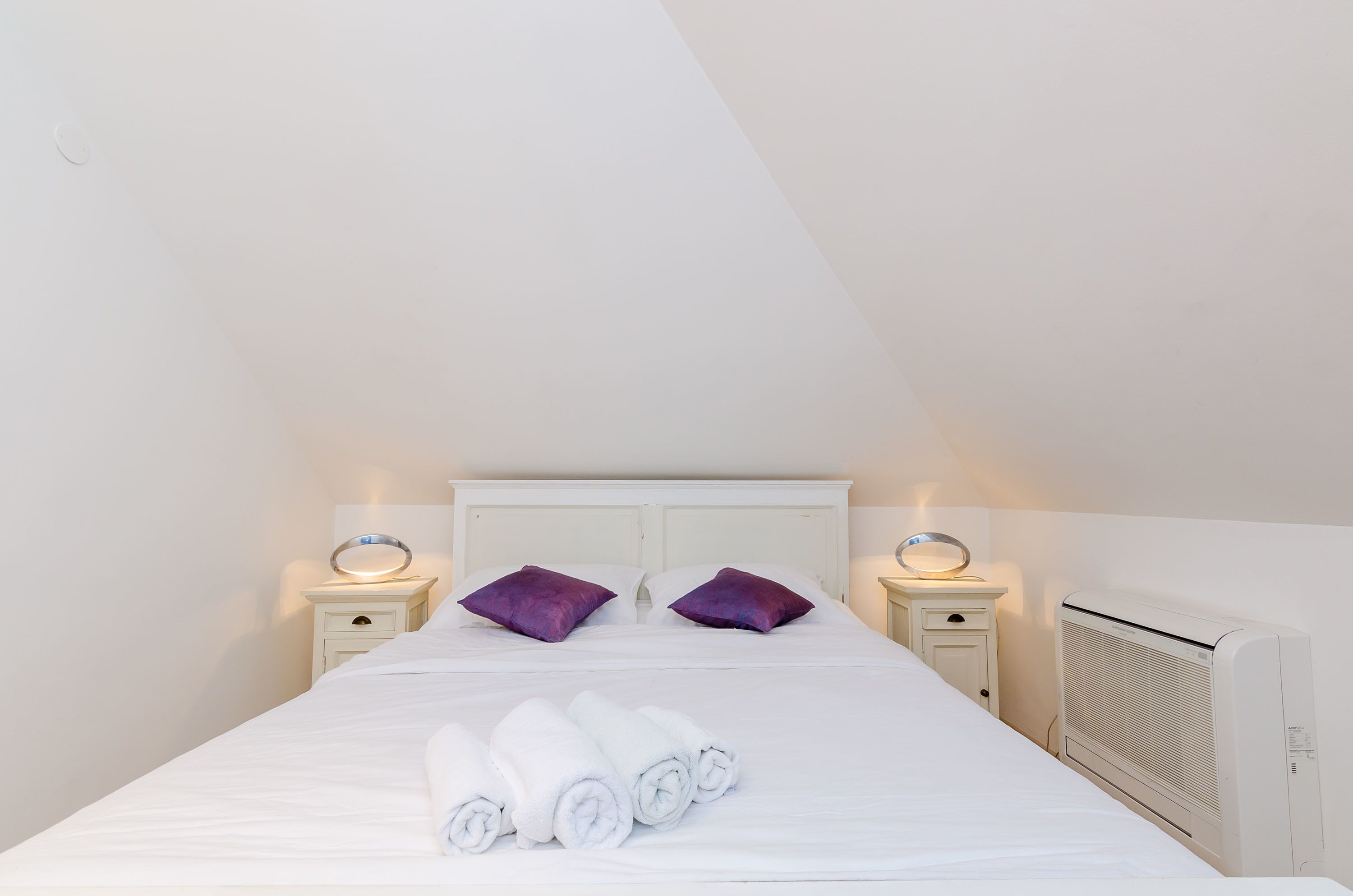A neatly made bed with white linens and rolled towels, two purple pillows, and a white headboard.