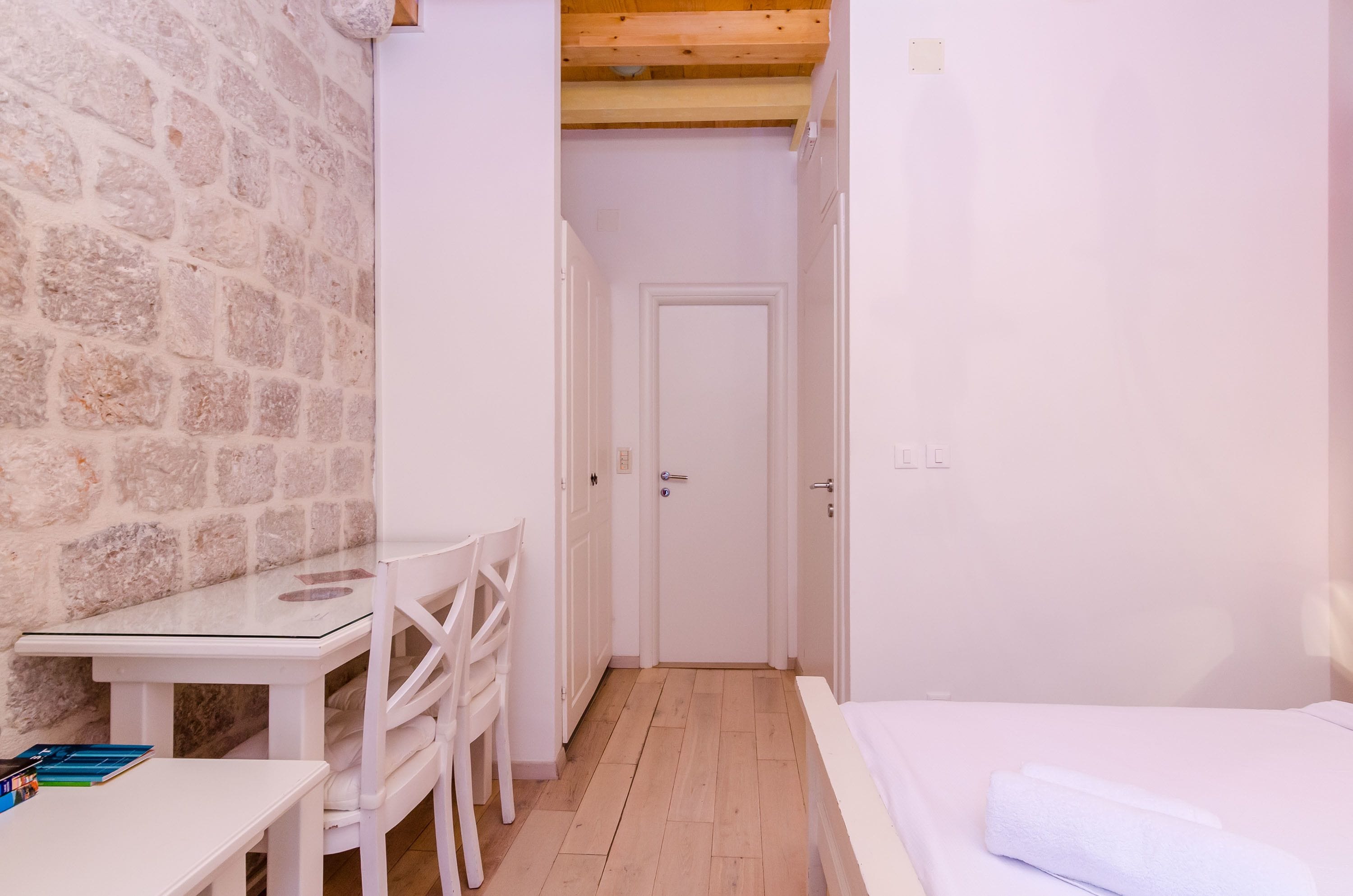 A narrow room with a stone wall, a wooden ceiling, a white door, a small table with chairs, and a bed with a white blanket.