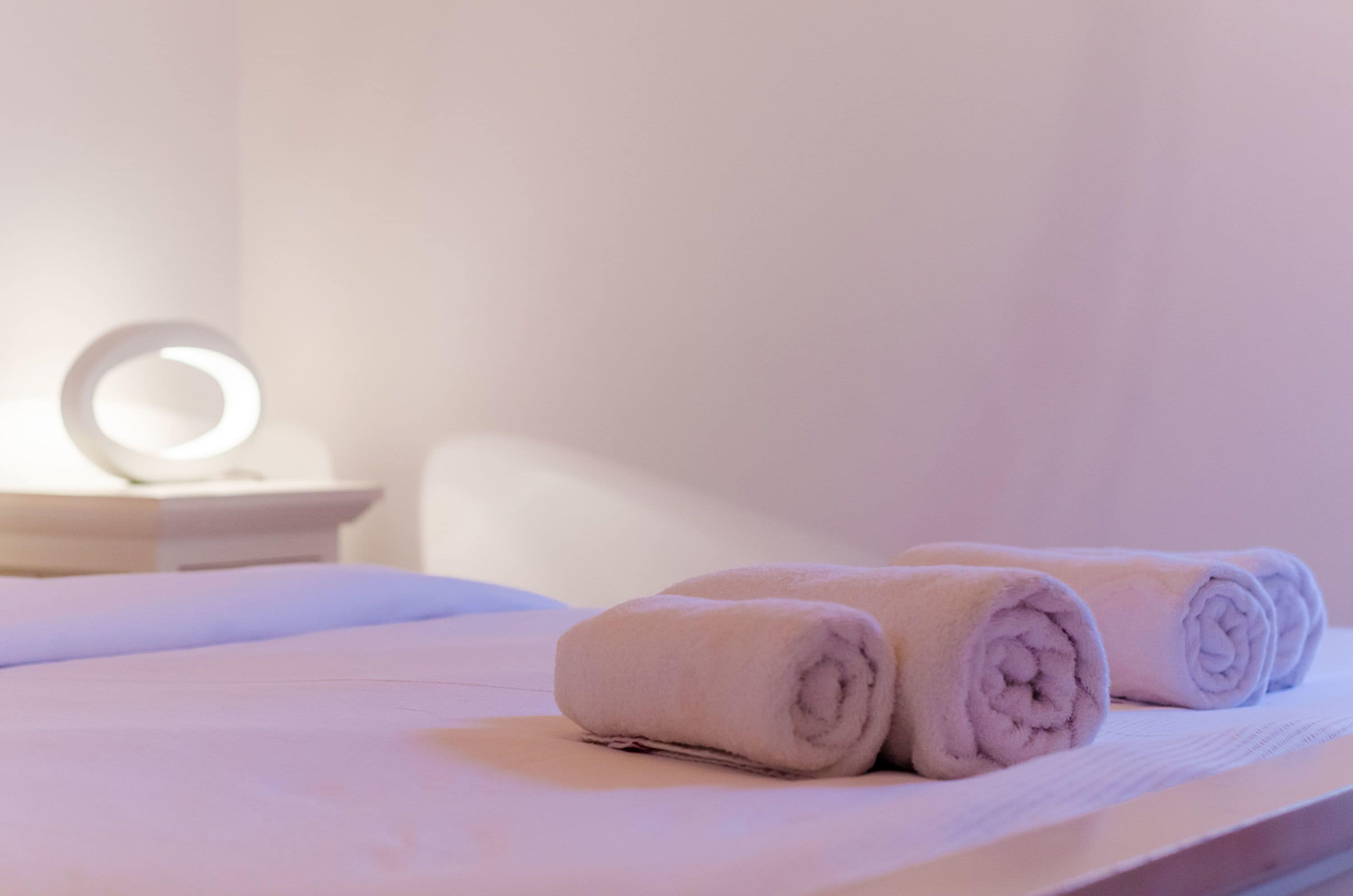 A neatly made bed with two rolled towels and a bedside table with a round mirror.